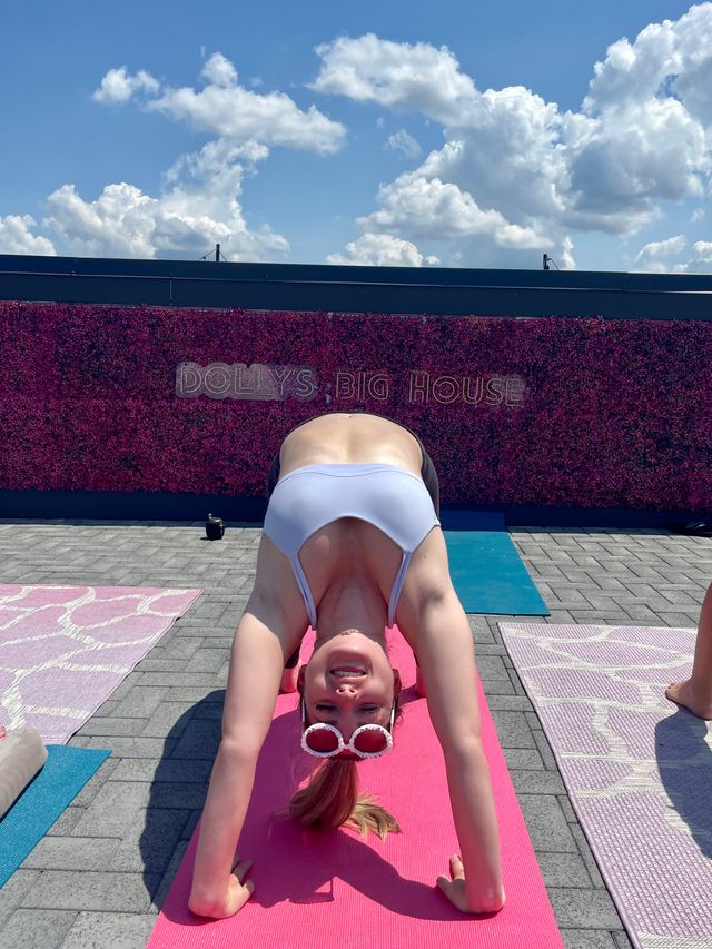 Person in white sports bra and round sunglasses in downward-dog on a pink yoga mat during sunny rooftop yoga in front of a pink floral wall under blue sky with fluffy clouds.