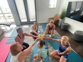 Six women on yoga mats in a bright apartment living room clinking glasses of orange juice in a cheerful post-workout toast, balcony doors and TV visible.