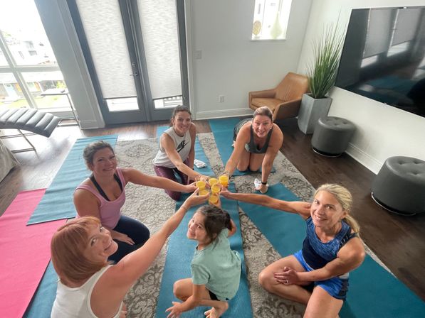 Six women on yoga mats in a bright apartment living room clinking glasses of orange juice in a cheerful post-workout toast, balcony doors and TV visible.