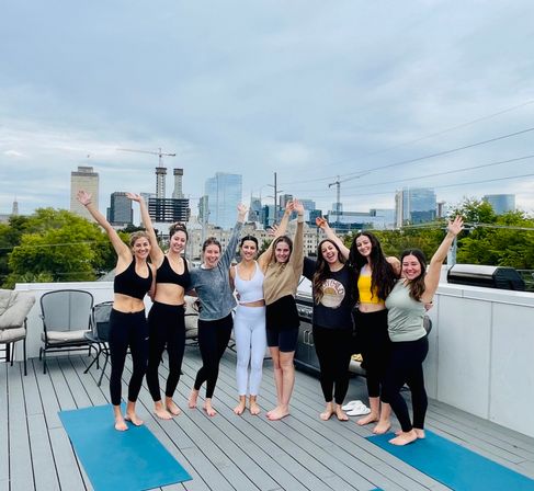 Eight friends barefoot on rooftop yoga mats, smiling with arms raised against a city skyline and construction cranes — outdoor rooftop yoga and wellness scene.