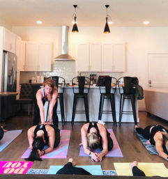 Small-group home yoga session in an open-concept modern white kitchen — instructor assists a student in child's pose on colorful mats while others rest, with bar stools, pendant lights and stainless appliances in the background.