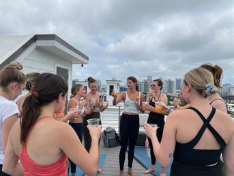 Women in activewear on a rooftop deck toasting with orange drinks after a yoga session, city skyline and cloudy sky in the background