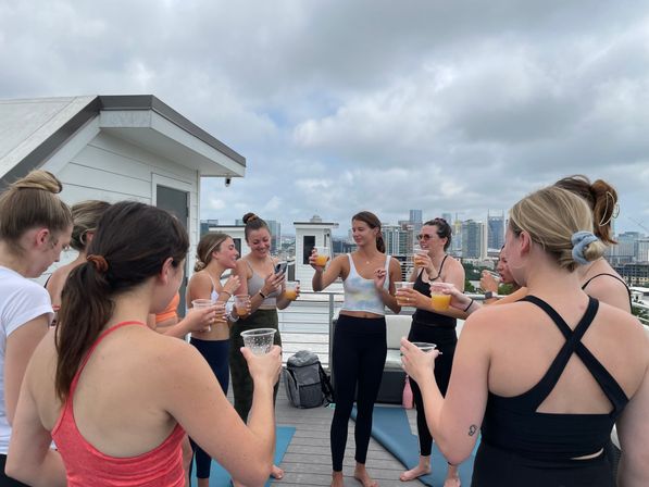 Women in activewear on a rooftop deck toasting with orange drinks after a yoga session, city skyline and cloudy sky in the background