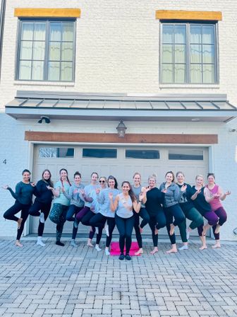 Group of women in leggings doing tree-pose yoga on a suburban white-brick house driveway in front of a garage, smiling together with the center participant wearing a sash.
