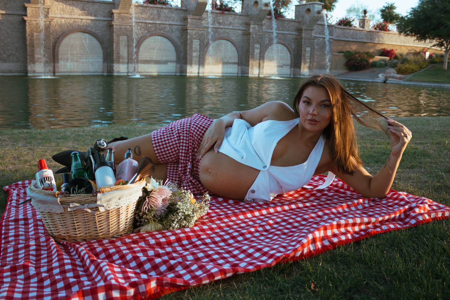Sunlit pregnant woman reclining on a red gingham picnic blanket by a pond with stone fountain arches, beside a picnic basket and flowers.