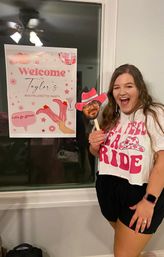 Smiling woman in a white and pink bachelorette T-shirt holding a pink cowboy-hat face prop beside a pink "Welcome" bachelorette party poster taped to a window.