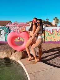Two friends in bikinis and heart-shaped sunglasses pose poolside beside a giant pink inflatable diamond-ring float, colorful mural wall and palm trees under a bright blue sky in a backyard setting.