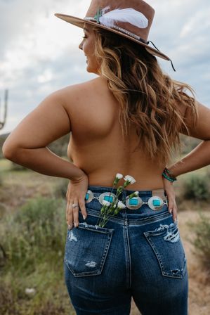Bare-backed woman wearing a feathered cowboy hat and turquoise concho belt, distressed denim jeans with white wildflowers tucked in the back pocket, standing in a sunlit Southwestern desert landscape.