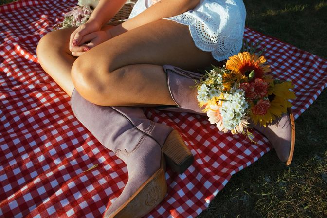 Sunlit outdoor picnic: crossed legs in a white eyelet dress, mauve suede cowboy boots on a red-and-white gingham blanket with a bright bouquet of sunflowers and daisies tucked into a boot