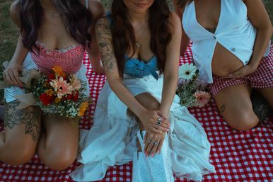 Three friends at an outdoor picnic on a red-and-white gingham blanket, holding floral bouquets; visible tattoos, center in a white dress with rings and a sparkly clutch.