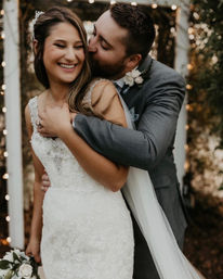 Smiling bride in lace wedding gown hugged and kissed on the cheek by groom in a gray suit beneath a twinkling outdoor arch with bouquet and string lights