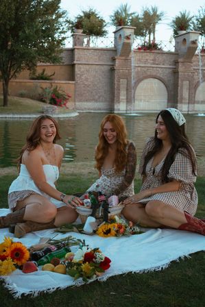 Three friends laughing at a lakeside picnic on a white blanket with flowers, fruit and drinks in a wicker basket in front of a decorative stone fountain wall