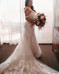 Bridal portrait in soft window light: woman in an off-the-shoulder lace wedding gown with a long floral train holding a rustic fall bouquet in a hotel room