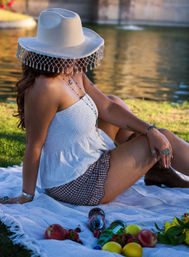 Lakeside picnic at golden hour: person lounging on a white blanket wearing a white beaded-fringe hat, smocked top, gingham shorts and boots, with apples, lemons, bottled drinks and sunflowers nearby.