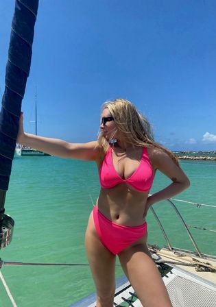 Person in a bright pink bikini and sunglasses posing on a sailboat deck with turquoise tropical water, clear blue sky, and distant sailboats — sunny beach getaway vibe.