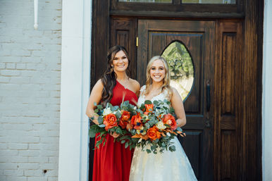 Smiling bride in a white lace gown and bridesmaid in a red dress holding vibrant orange-rose and eucalyptus bouquets in front of a dark wooden entry door.