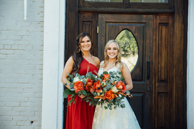 Smiling bride in a white lace gown and bridesmaid in a red dress holding vibrant orange-rose and eucalyptus bouquets in front of a dark wooden entry door.