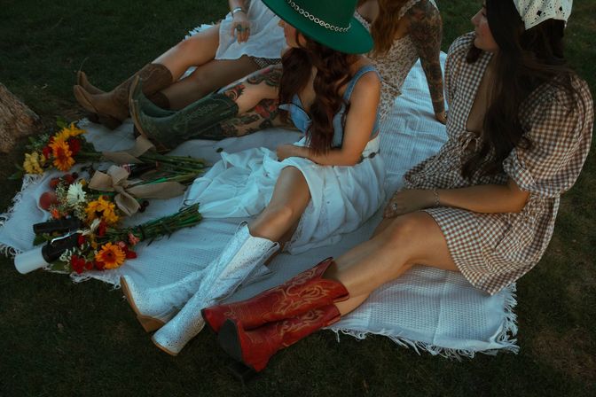 Three women at a boho picnic on a grassy lawn, seated on a white blanket wearing colorful cowboy boots and sundresses, one in a green hat with sunflowers and wildflower bouquets nearby.