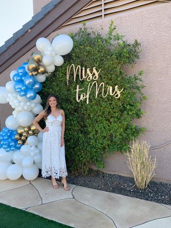 Smiling woman in a white lace dress at an outdoor patio bridal shower, posing by a leafy green hedge with a wooden “Miss to Mrs” sign and a white, blue and gold balloon arch.