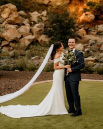 Bride in an off-the-shoulder white gown with long veil and groom in a dark suit smiling at an outdoor wedding by a sunset-lit rocky hillside, holding a leafy bouquet.