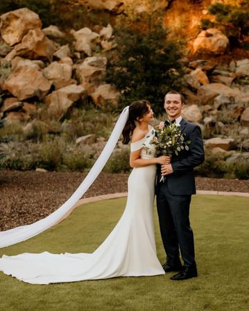 Bride in an off-the-shoulder white gown with long veil and groom in a dark suit smiling at an outdoor wedding by a sunset-lit rocky hillside, holding a leafy bouquet.
