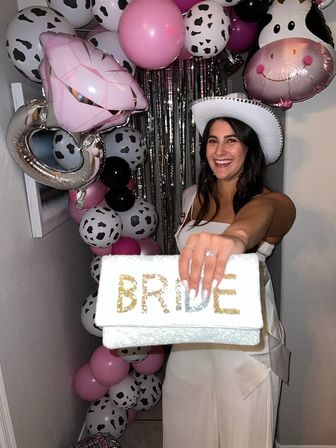 Smiling woman in a white cowgirl hat at an indoor bachelorette party holding a glittery clutch labeled “BRIDE” in front of pink, cow-print and metallic balloons.