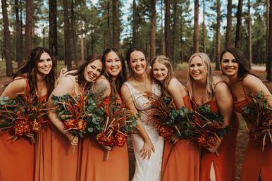 Smiling bride in a white lace gown surrounded by six bridesmaids in burnt-orange dresses holding eucalyptus and orange bouquets in a pine forest outdoor wedding portrait.