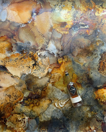Overhead view of an abstract marbled surface in rust, gold, amber and gray tones with a tilted whiskey bottle resting on the textured background