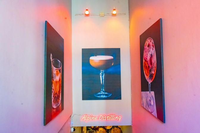 Vibrant pink-lit staircase leading to a bar entrance, flanked by three oversized cocktail paintings with overhead spotlights and a glowing pink neon sign.