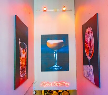 Vibrant pink-lit staircase leading to a bar entrance, flanked by three oversized cocktail paintings with overhead spotlights and a glowing pink neon sign.