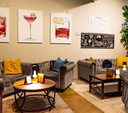 Cozy indoor lounge with tufted gray sofas, yellow and blue accent pillows, wooden coffee tables, leafy potted tree and colorful cocktail canvas art on the wall.