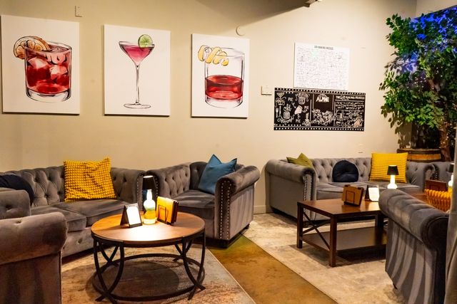 Cozy indoor lounge with tufted gray sofas, yellow and blue accent pillows, wooden coffee tables, leafy potted tree and colorful cocktail canvas art on the wall.