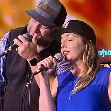 Close-up of two singers performing a live duet on stage — a bearded man and a woman both wearing caps and holding microphones against a warm orange backdrop.