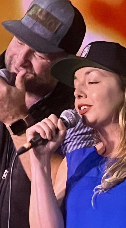Close-up of two singers performing a live duet on stage — a bearded man and a woman both wearing caps and holding microphones against a warm orange backdrop.