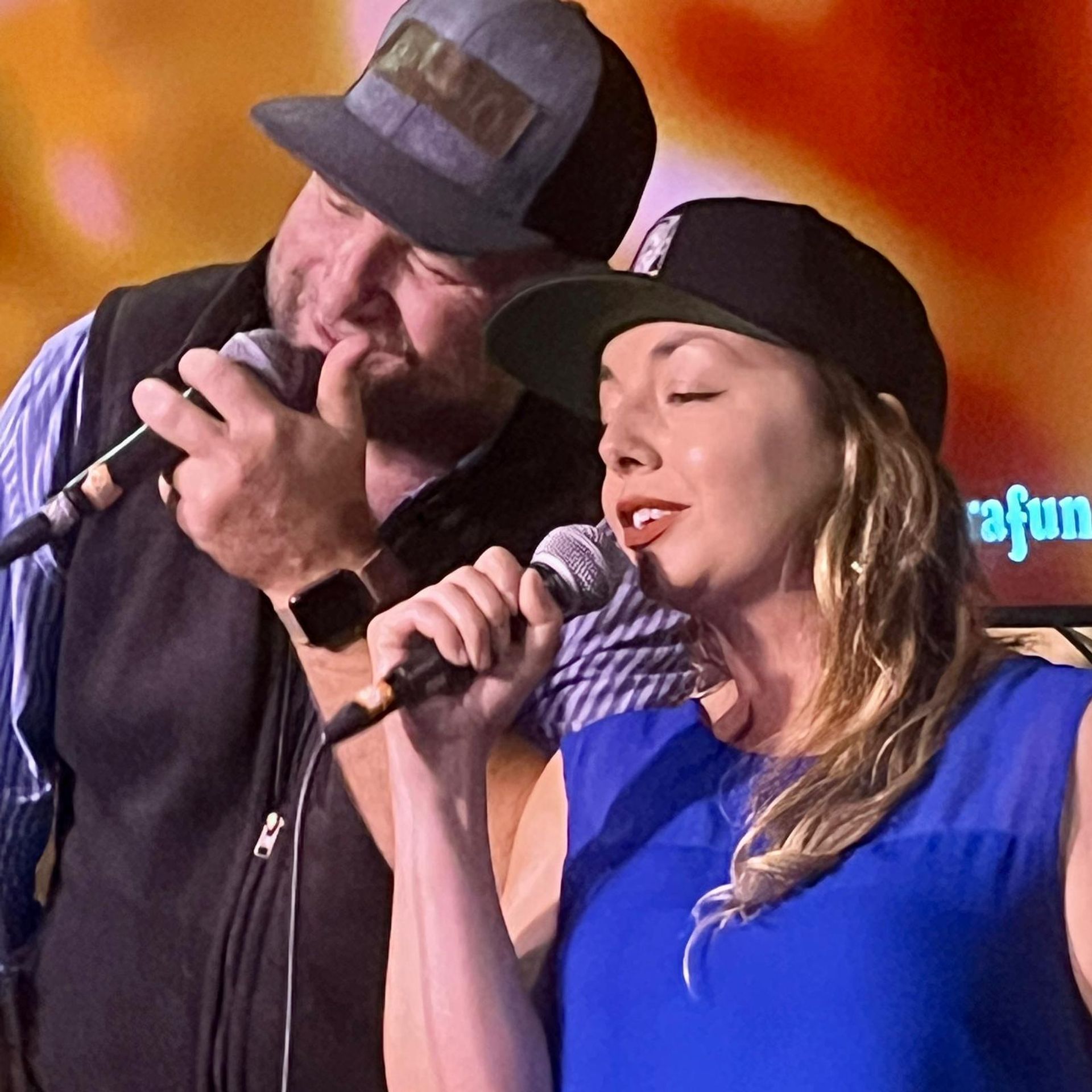 Close-up of two singers performing a live duet on stage — a bearded man and a woman both wearing caps and holding microphones against a warm orange backdrop.