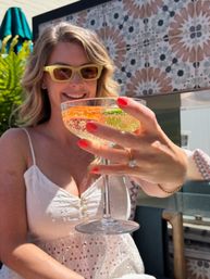 Smiling woman in yellow sunglasses on a sunny outdoor tiled patio holding a champagne coupe garnished with orange and lime, showing red nails, a diamond ring and white eyelet dress.