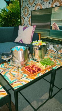 Vibrant mosaic-tiled patio table set for cocktails with metal ice buckets of canned drinks, coupe glasses, and a wooden board of blood orange slices, lime wedges and fresh herbs beside a blue sofa and patterned tile wall.