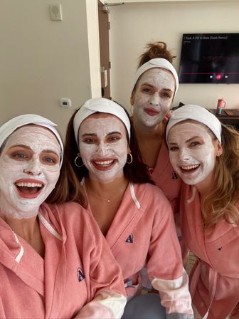 Four friends in matching pink robes and white headbands wearing white facial masks, smiling for a selfie during a spa day in a hotel room.