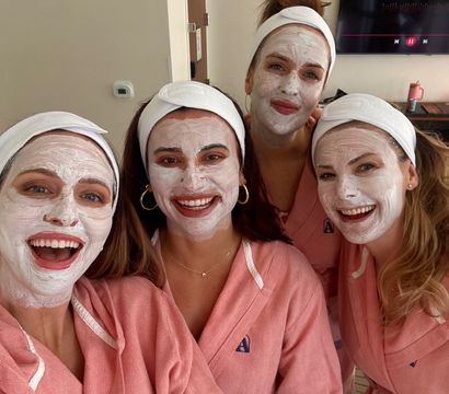 Four friends in matching pink robes and white headbands wearing white facial masks, smiling for a selfie during a spa day in a hotel room.