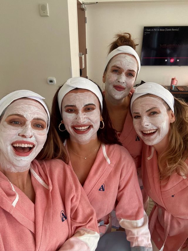 Four friends in matching pink robes and white headbands wearing white facial masks, smiling for a selfie during a spa day in a hotel room.