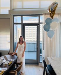 Smiling woman in a white sundress poses in a bright modern downtown hotel suite by glass doors to an urban balcony, with blue-and-white balloons floating beside a set dining table.