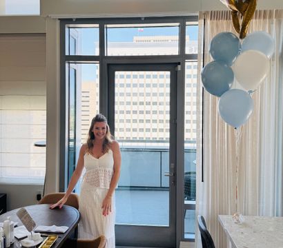 Smiling woman in a white sundress poses in a bright modern downtown hotel suite by glass doors to an urban balcony, with blue-and-white balloons floating beside a set dining table.