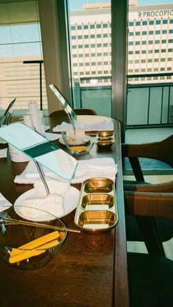 Sunlit balcony manicure station on wooden table with hand mirrors, gold trays, bowls, cotton towels and yellow nail tools, city high-rise view