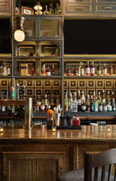 Cozy upscale cocktail bar interior — wood counter with brass taps, lit candles, and backlit shelves of glinting whiskey and liquor bottles.