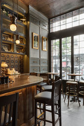 Cozy vintage-style bar interior with a curved wooden counter, leather high stools, brass pendant lights, decorative shelving and candles, and tall paned windows overlooking a city street.