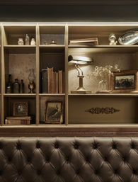 Cozy vintage study shelf with wooden cubby shelves holding antique books, glass bottles, framed photos, a banker's lamp and dried flowers above a tufted leather bench