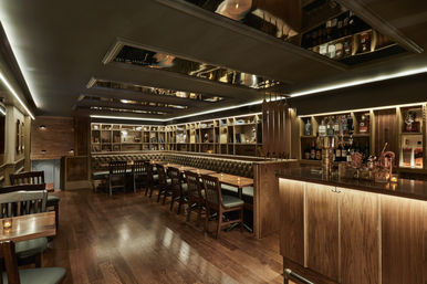 Speakeasy-style cocktail lounge interior — dimly lit with warm wood floors and bar, tufted leather banquette, long communal tables, backlit bottle shelving and mirrored ceiling panels.