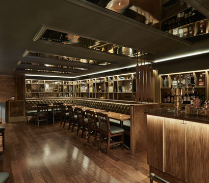 Speakeasy-style cocktail lounge interior — dimly lit with warm wood floors and bar, tufted leather banquette, long communal tables, backlit bottle shelving and mirrored ceiling panels.