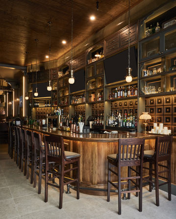 Inviting upscale wooden cocktail bar interior with a curved polished counter, row of high wooden stools, backlit shelves of liquor bottles and glassware, vintage cabinetry, hanging globe pendant lights and warm amber ambiance.