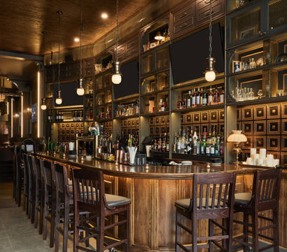 Inviting upscale wooden cocktail bar interior with a curved polished counter, row of high wooden stools, backlit shelves of liquor bottles and glassware, vintage cabinetry, hanging globe pendant lights and warm amber ambiance.